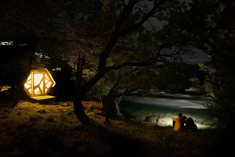 Un refugio encendido de noche junto al río Cochrane, con una pareja frente al agua