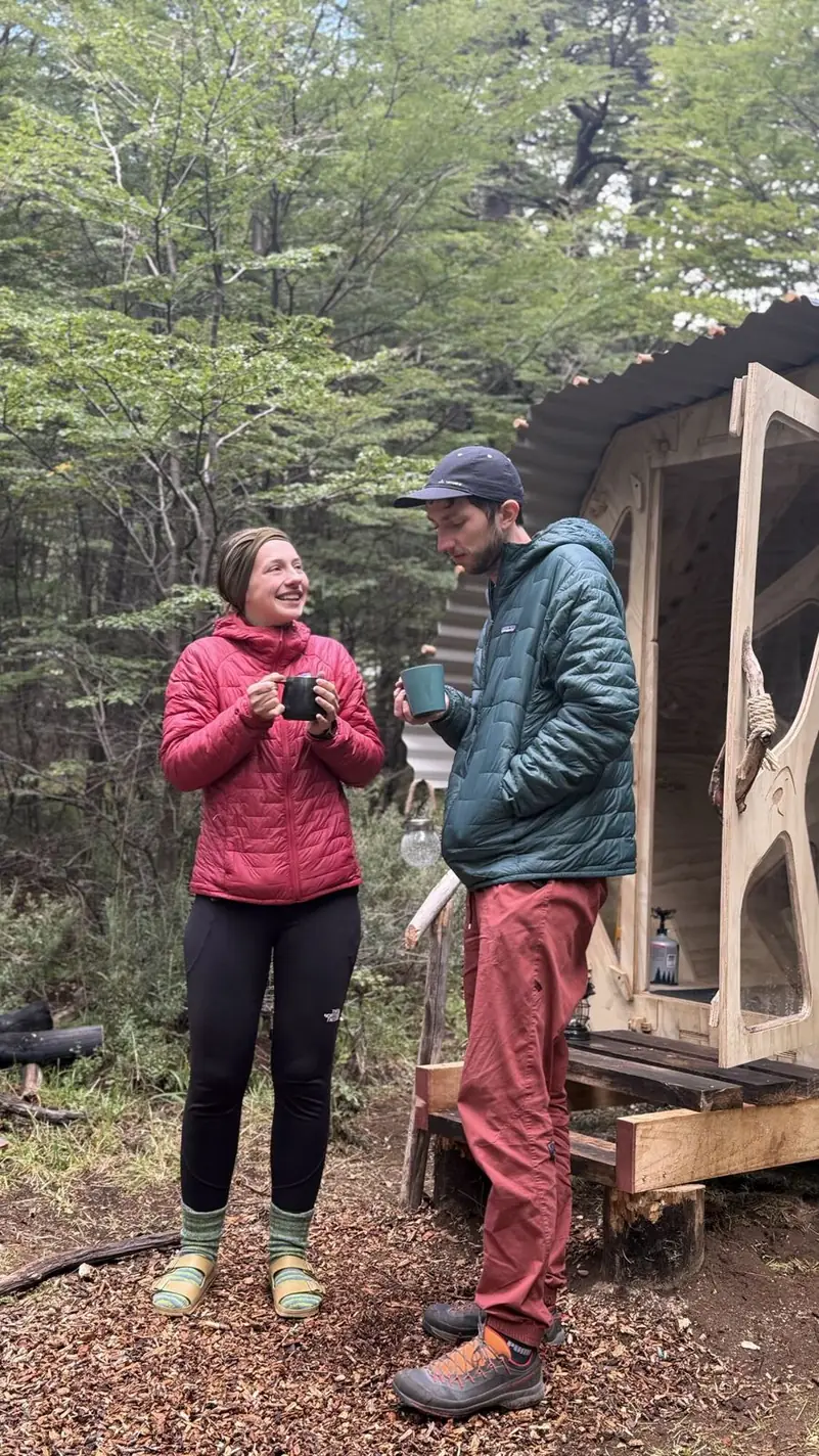 Dos personas conversan con un café junto al refugio, en el bosque